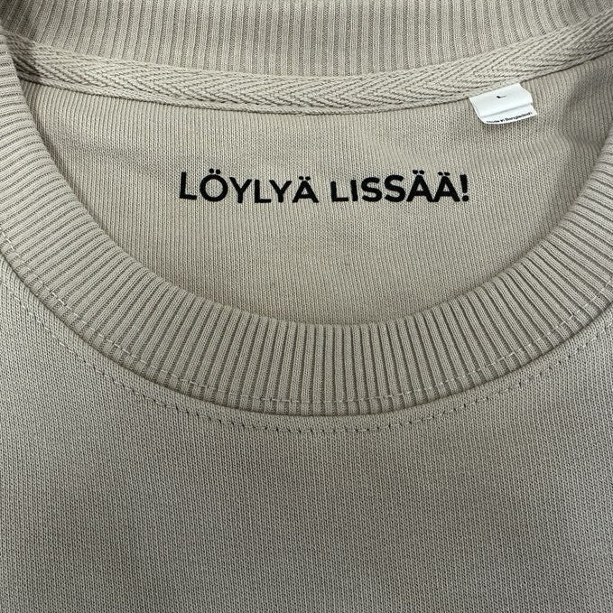 Close-up of a ribbed crewneck sweatshirt in a light beige or oatmeal color, featuring the black printed text 'LÖYLYÄ LISSÄÄ!' on the inner neck label.