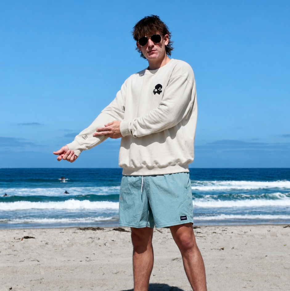A man wearing a cream sweatshirt and light blue shorts stands on a sandy beach with the ocean and blue sky in the background. He is wearing sunglasses and stretching his arm.