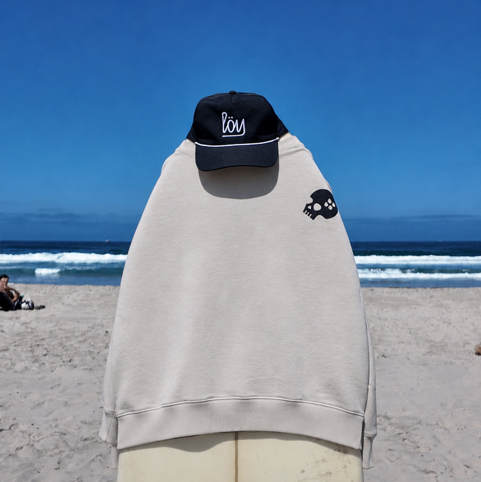 A light beige hoodie with a small black skull graphic on the left chest, draped over a mannequin on a sandy beach with the ocean and blue sky in the background. A black baseball cap with white script is placed on top of the hoodie.
