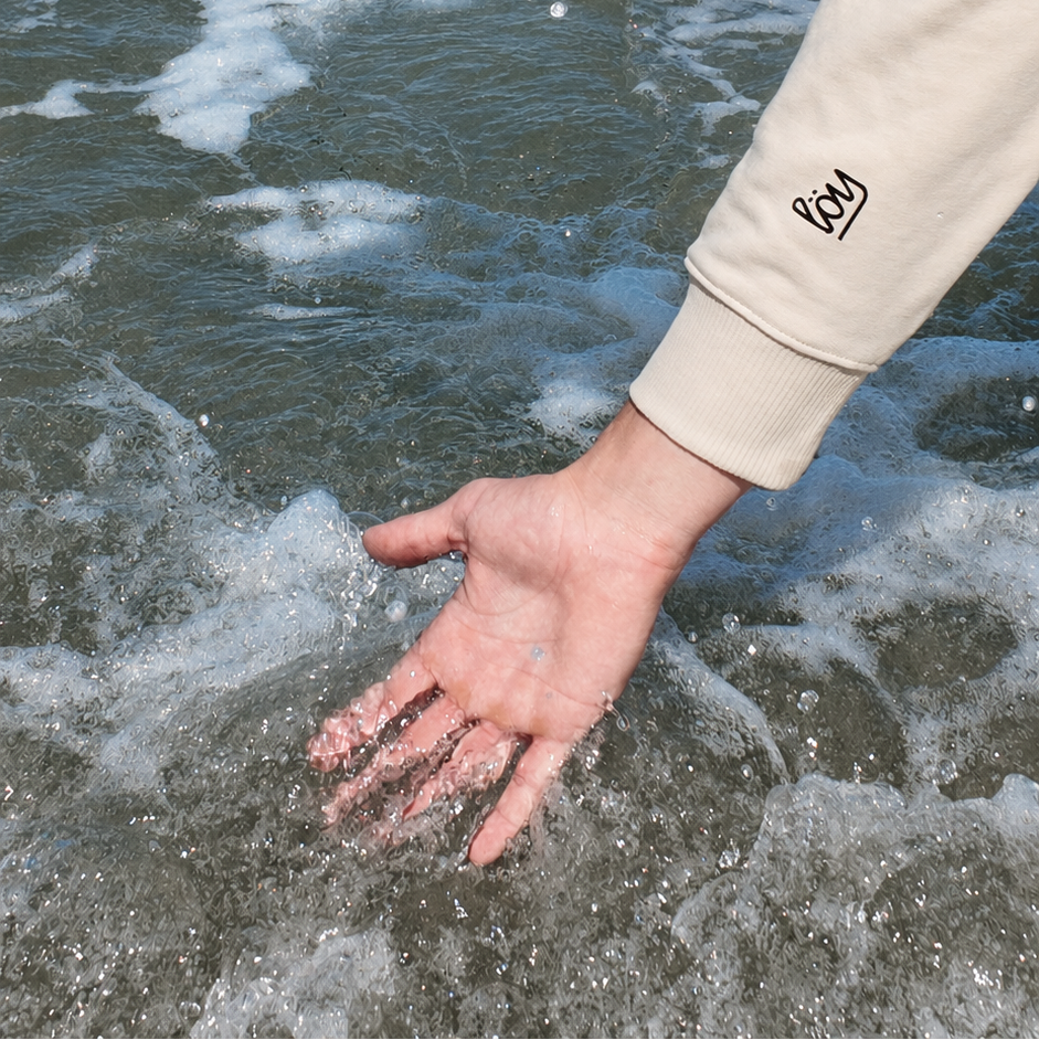 A close-up of a person's hand reaching into foamy ocean water, with their beige sleeve visible on the right side. The sleeve features a small black embroidered logo that reads 'PDA'.