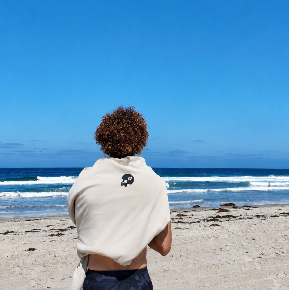 A person standing on a sandy beach with their back to the camera, wearing a cream-colored hoodie with a small black skull logo on the back, looking out toward the ocean under a clear blue sky.